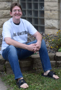 Photo of 30-something, Caucasian male, Chris, sitting outside in a white LIVE UNITED t-shirt and jeans