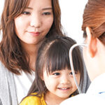 Young Asian girl sitting on mother's lap while female doctor listens to her heart with a stethoscope