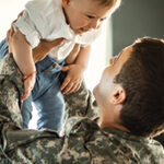 Caucasian man in military camouflage lifting a small baby up over his head